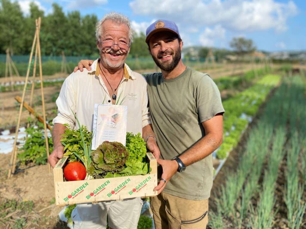 Maraîchers Bio et Légumes Frais : Le Goût d'Eden en Direct Deux maraîchers souriants avec une cagette BIOGARDEN remplie de légumes frais dans leur potager bio en plein air.